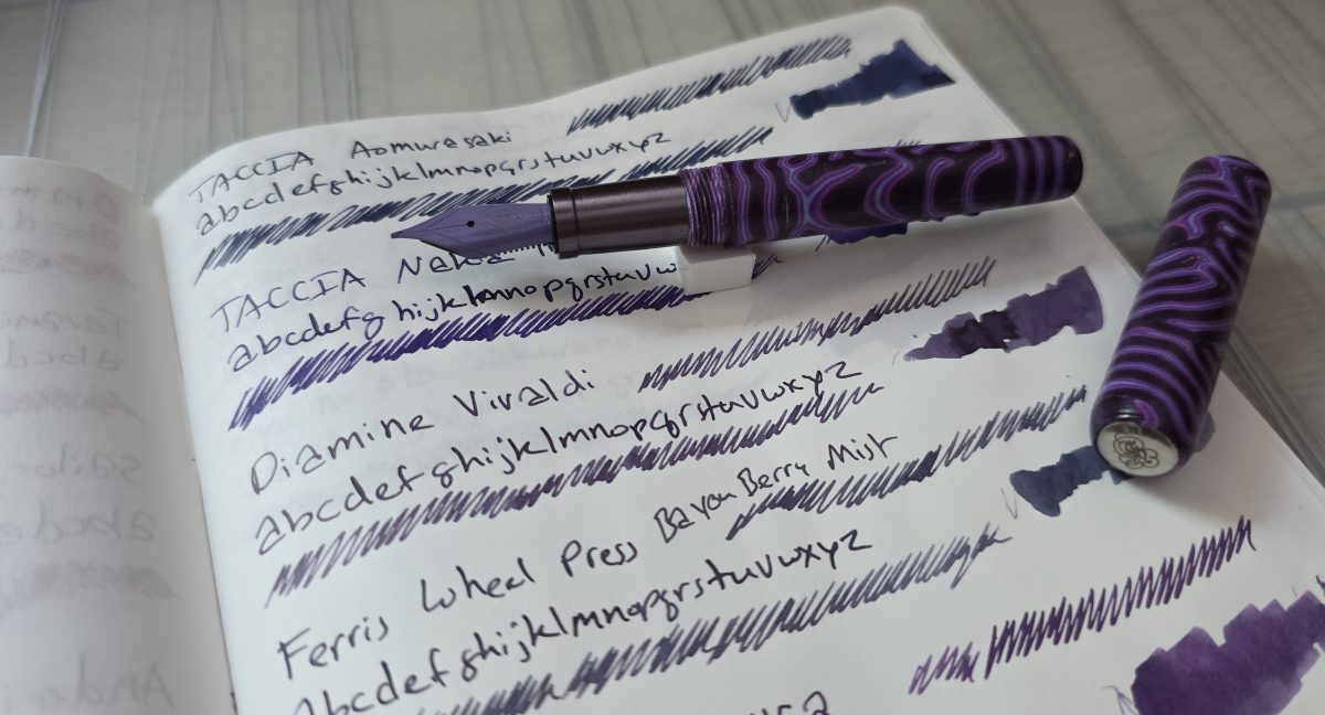 A swatch and writing sample page of multiple dark purple inks with the dark, dusty purple Diamine Vivaldi in focus, and a Crazyfiber purple micarta pocket pen uncapped with a lilac nib and dark gray purple metal section resting on the page.