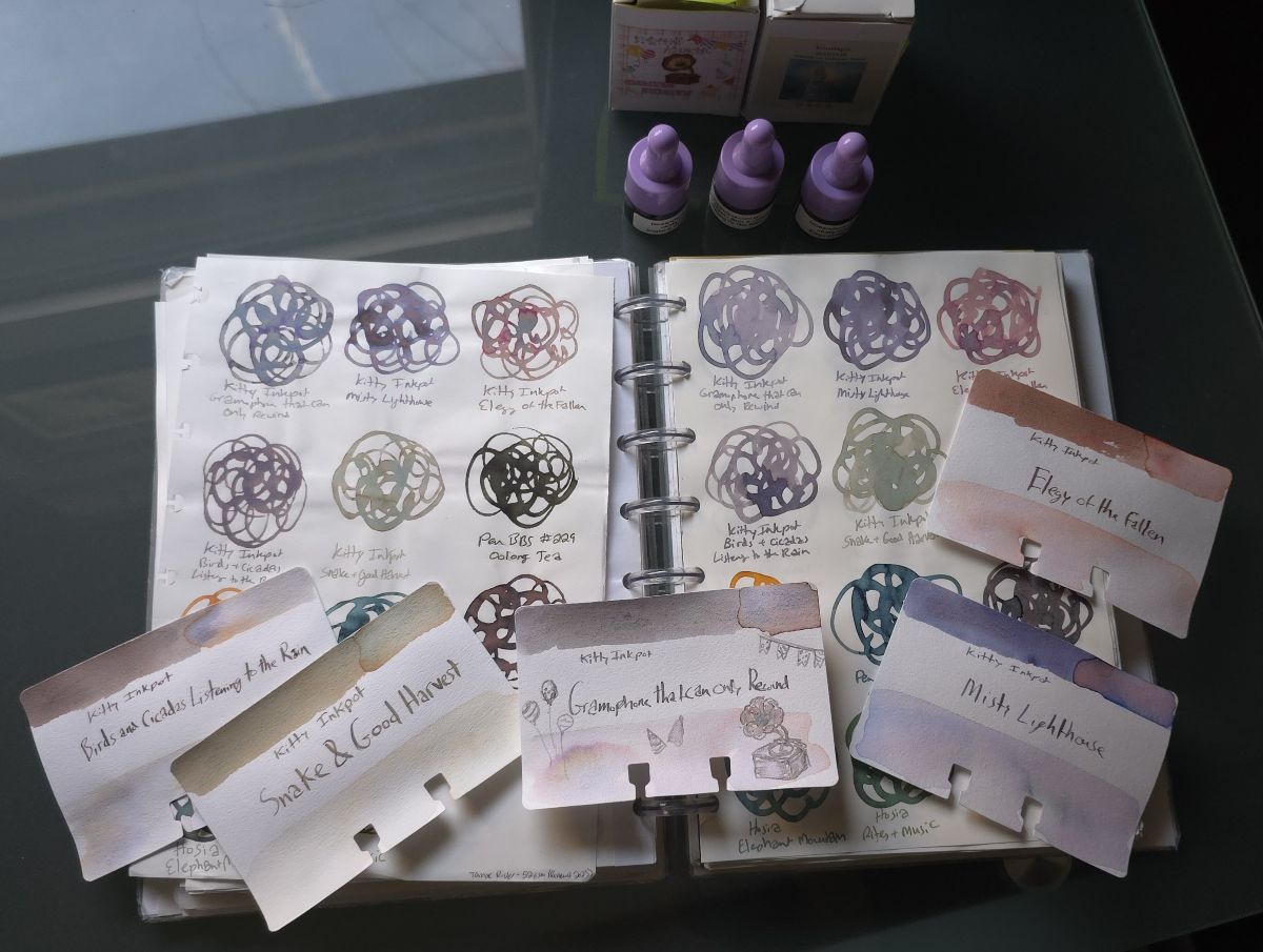 A disc-bound swatchbook open to two pages of swatches of chromashading inks from Kitty Inkpot, with the related bottles and samples of ink and five Col-o-dex swatch cards for Birds and Cicadas Listening in the Rain, Snake and Good Harvest, Gramophone That Can Only Rewind, Misty Lighthouse, and Elegy of the Fallen.