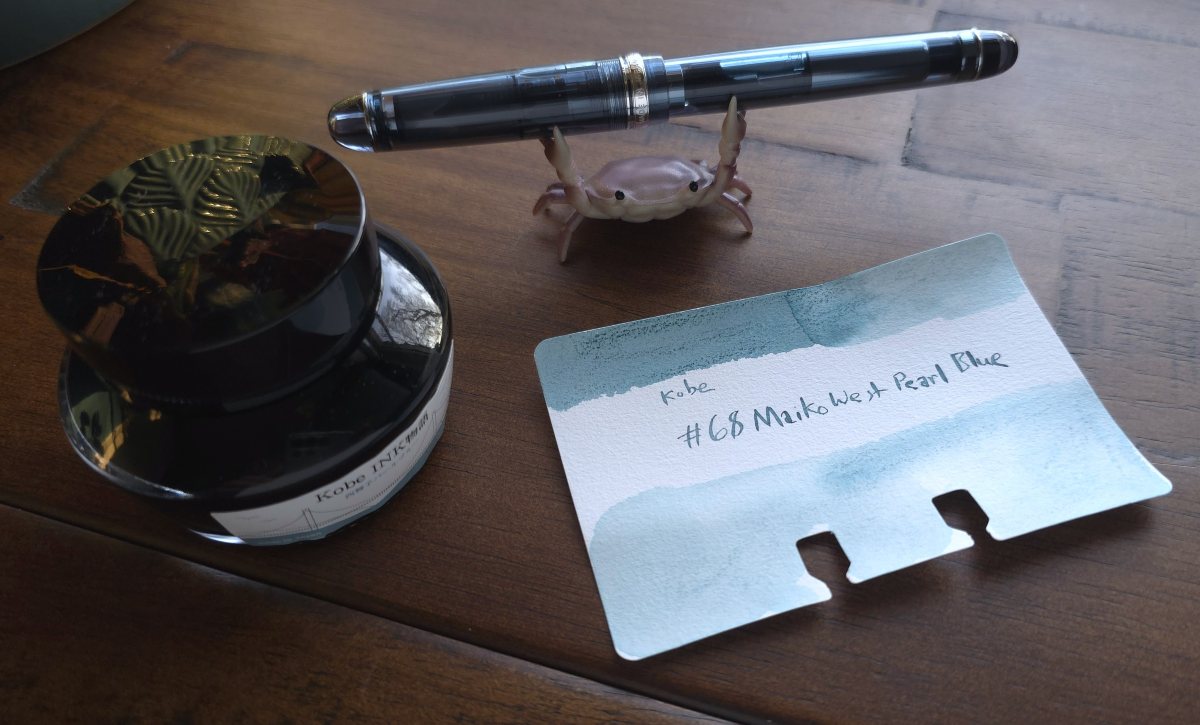 A Col-o-dex ink swatch card for the light blue gray Kobe #68 Nishimaiko (or Maiko West) Pearl Blue ink, next to the glass ink bottle and a Pilot 74 fountain pen in a similar gray blue shade.