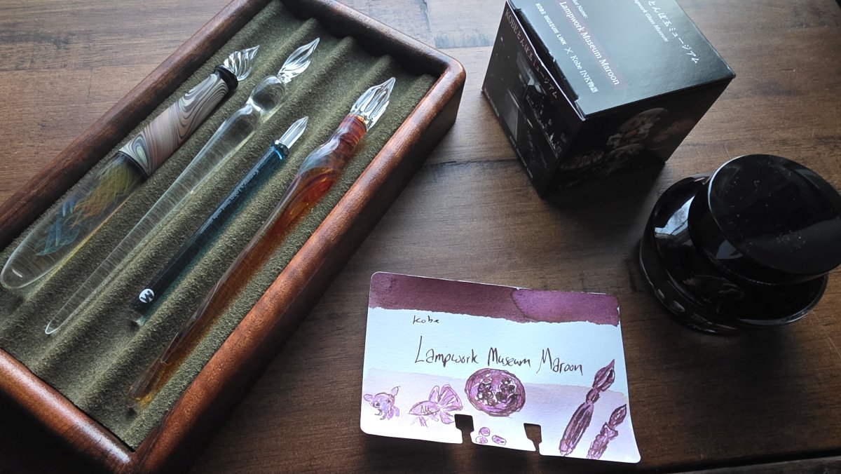 Pen tray with four nice glass dip pens next to a swatch card with a sketch and the ink bottle and box for Kobe Lampwork Museum Maroon.