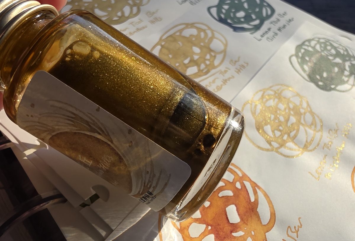 A glass bottle of golden brown fountain pen ink with shimmer in the sun with swirled swatches out of focus in the background.