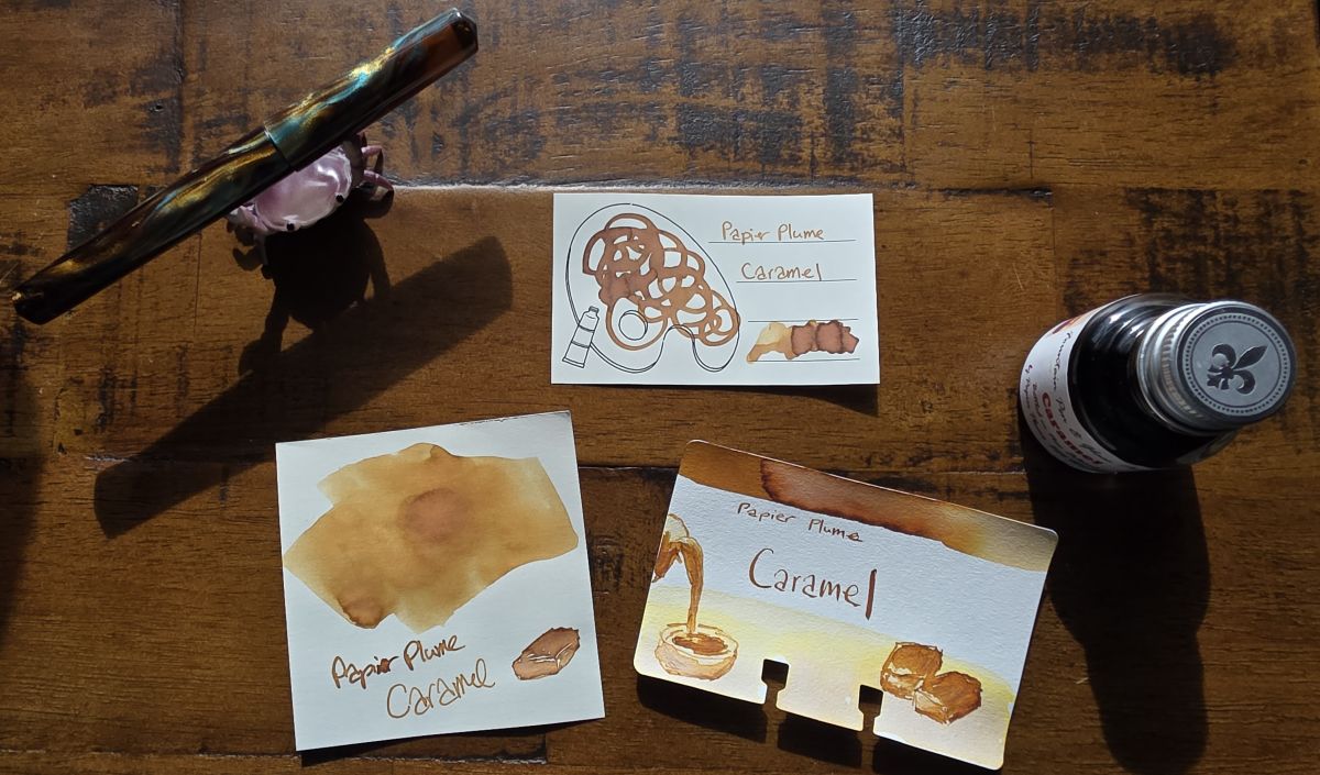 A trio of ink swatches on different paper for the golden yellow brown Papier Plume Caramel ink, with the signature bottle and an acrylic fountain pen with brown, light brown, and blue swirled material.