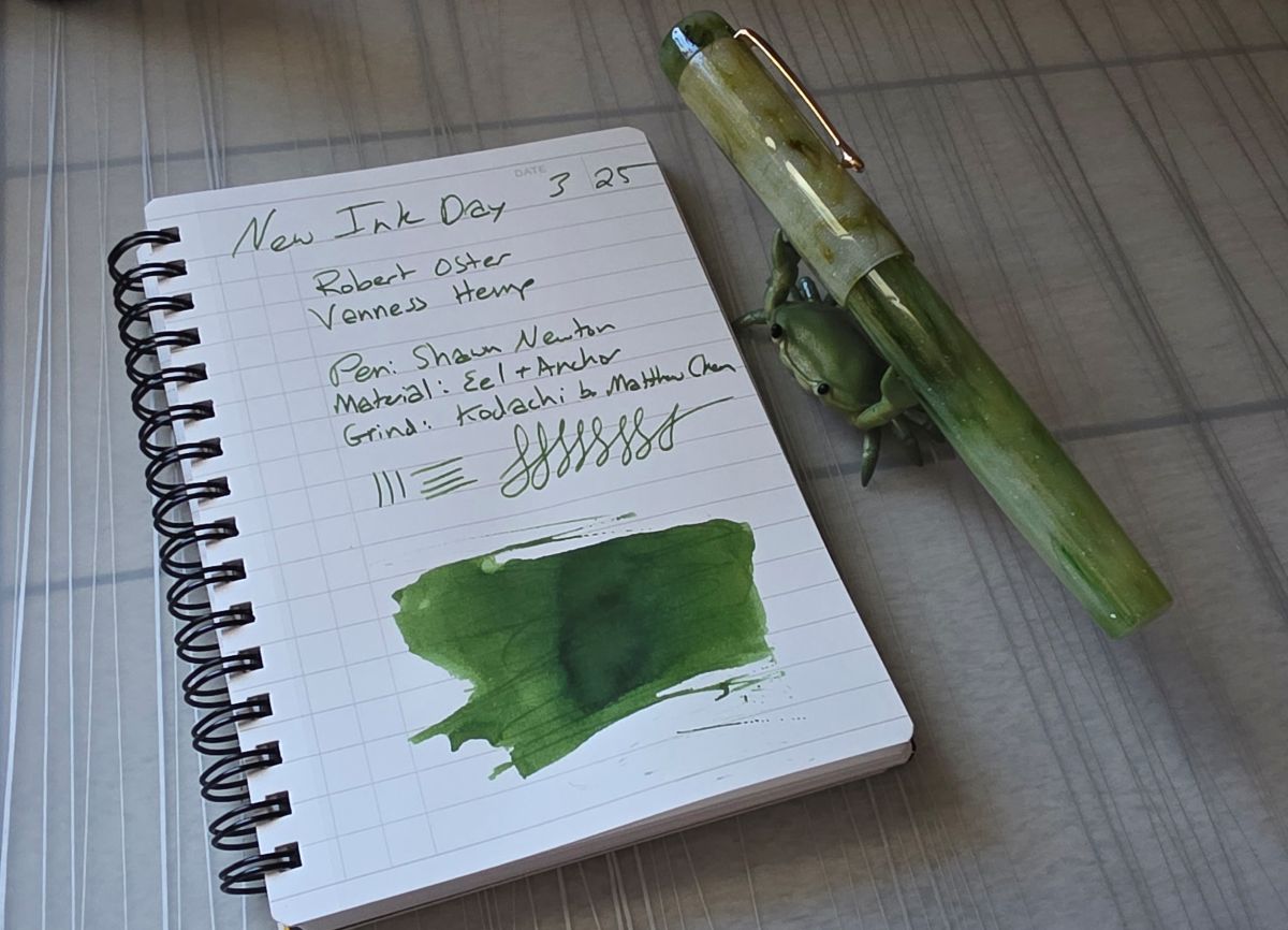 A spiral notebook with writing and ink swatch for a rich green ink called Robert Oster Vanness Hemp, next to a turned resin fountain pen in swirled green and white being held by a green crab pen rest.