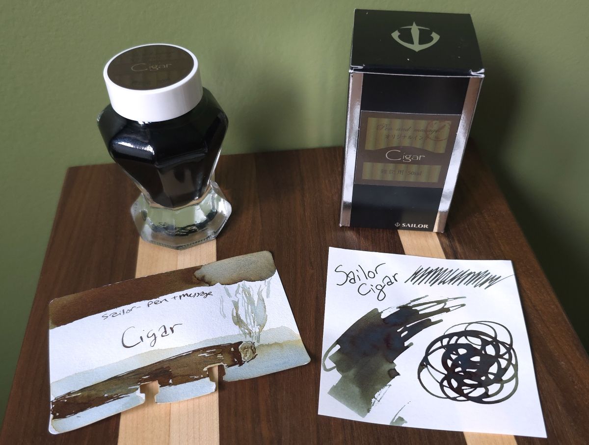 Sailor Cigar fountain pen ink in a vase bottle with the box and a couple ink swatches including a Col-o-dex card with a sketch of a cigar and a lot of rusty sheen and Graphilo paper where the ink is more blue green.