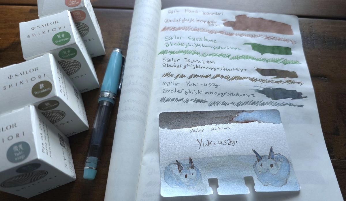 A swatch page for four Sailor Shikiori inks in dusty pink, leafy green, gray brown, and gray blue, with the corresponding ink bottles, and a Col-o-dex swatch card with a sketch of snow bunnies.