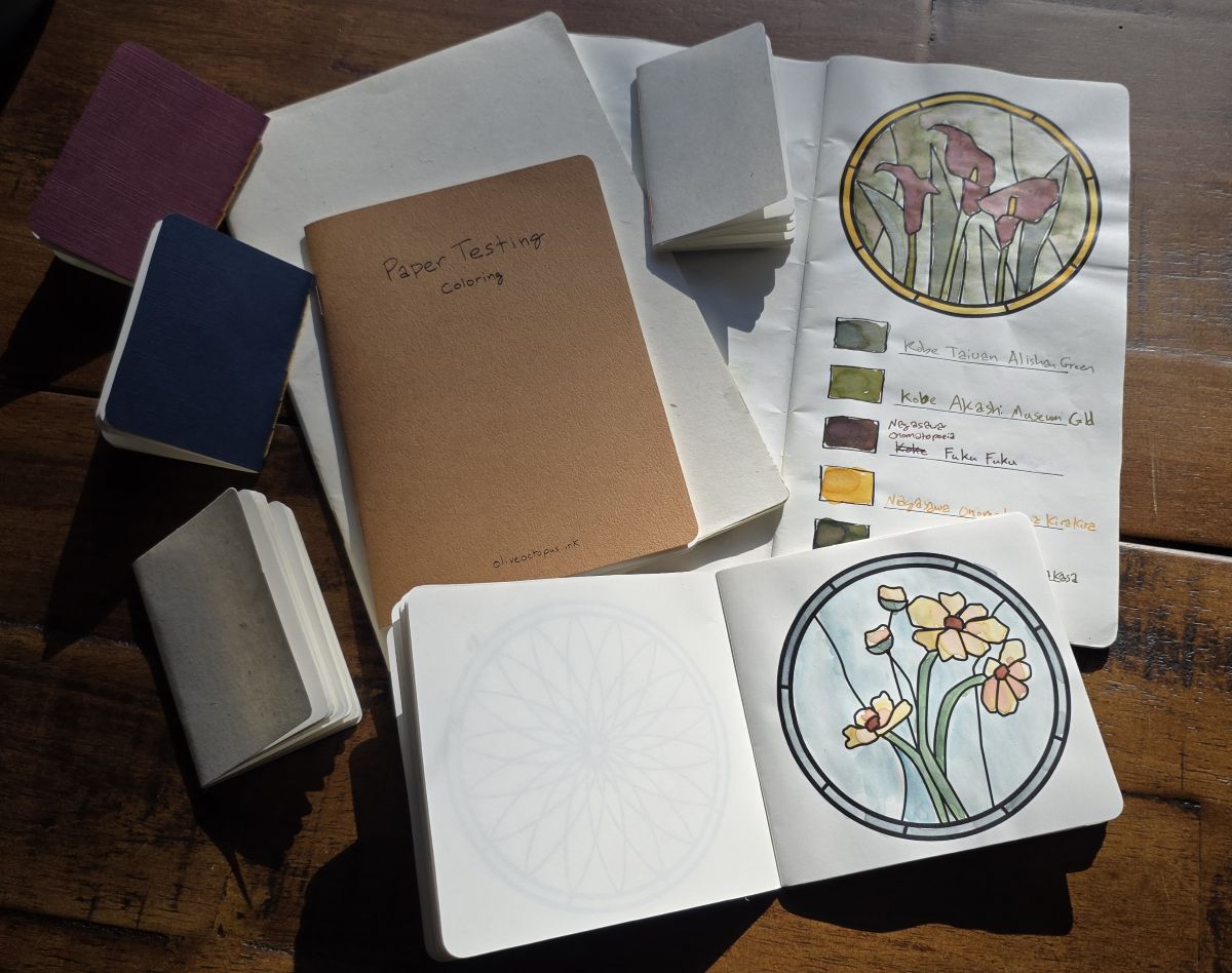 A collection of hand-made booklets including an A5 slim booklet with a stained glass flower outline and swatches painted in, a small square coloring book with a different stained glass outline, an A6 paper testing booklet, an A5 booklet, and several small paper booklets.