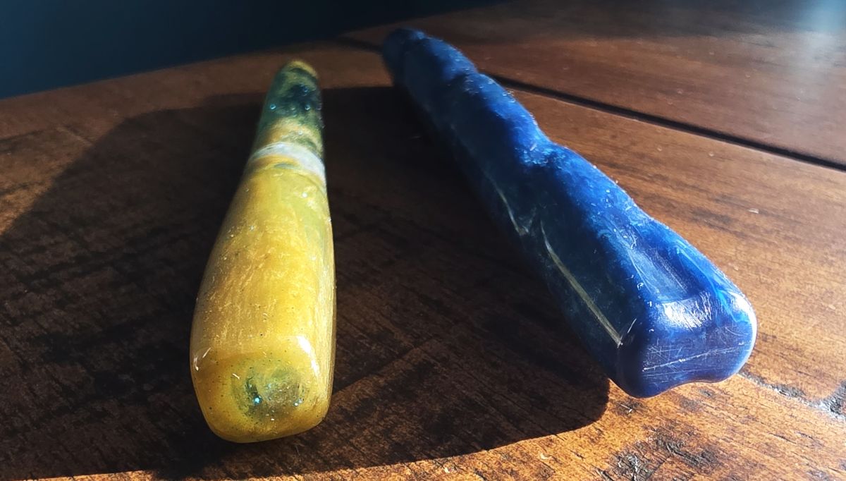 The ends of the caps of two hand-carved fountain pens, a green-gold resin pen with a squared cap and a sparkly medium blue resin pen with a carved spiral and twist along the whole pen body.