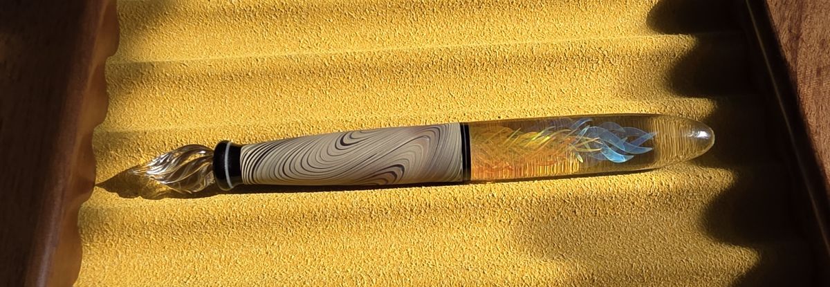 A yellow pen tray holding a handmade glass dip pen with a matte section that looks like wood grain and a clear body with artistic swirls of blue and yellow at the center.