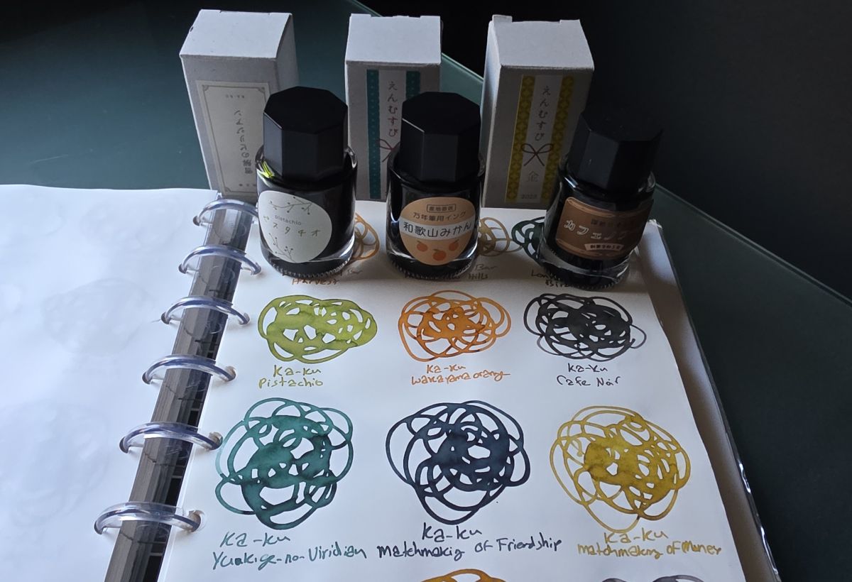 A sheet of paper in a disc-bound notebook with swatches of six colors of Ka-Ku fountain pen inks and the corresponding ink bottles, including Pistachio, Wakayama Orange, Cafe Noir, Yukige-no-Viridian, Matchmaking of Friendship, and Matchmaking of Money.