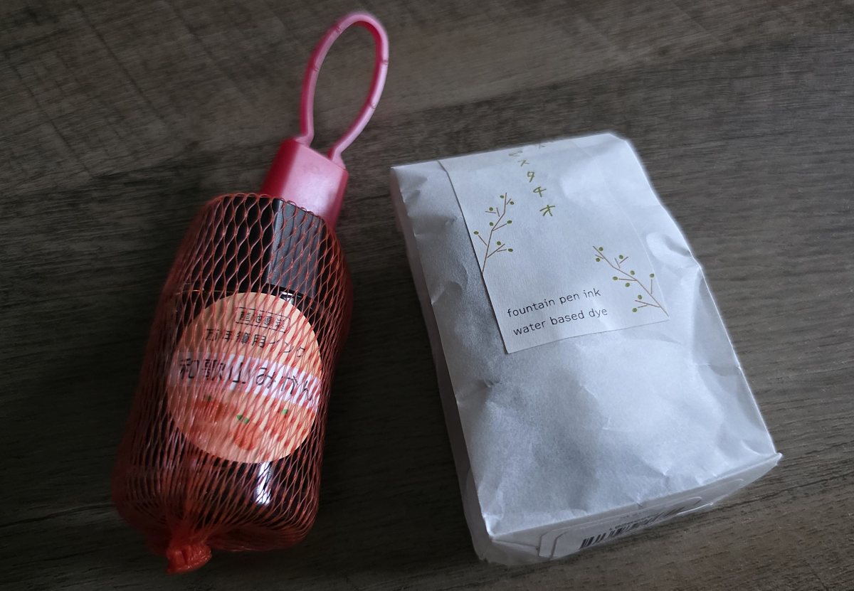 Two glass bottles of fountain pen ink, one in a tiny orange net bag like that used to hold oranges at the grocery store, and the other in a white tracing paper bag sealed with a printed label sticker.