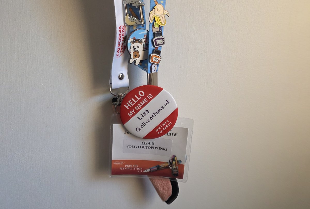 A pen show lanyard covered in enamel pins and a red and white button that says 'Hello my name is Lisa @oliveoctopus.ink and I am a Pen Addict!' holding a name tag for the DC Pen Supershow and a fuzzy peach single Rickshaw pen sleeve, and a second lanyard from the California Pen Show.