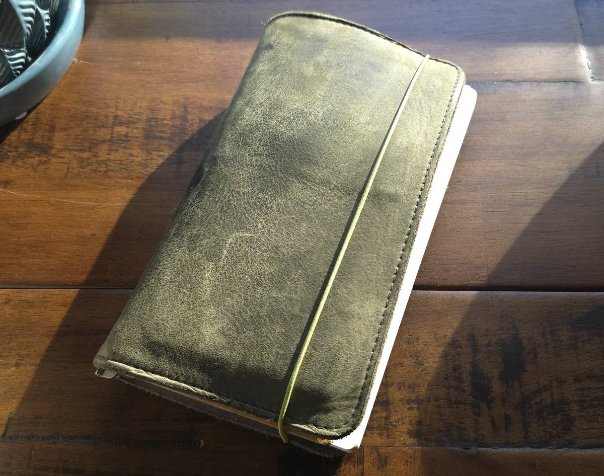 A TGS_25 A5 Slim Roterfaden Taschenbegleiter leather notebook cover in olive green with a light green band holding it closed, sitting on a wood desk in afternoon sunlight.