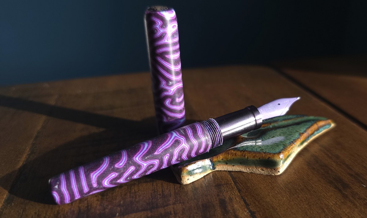 An uncapped pocket fountain pen made of a wild purple patterned micarta with a dark gray purple metal section and a lilac color fountain pen nib.