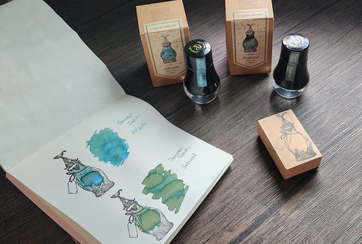 A notebook with two stamped fancy bottle designs and ink swatches for two Dominant Industry inks, a blue green ink with bright blue shimmer called Atlantis and a shading green called Seaweed, plus the bottles for the two inks and the wooden stamp.