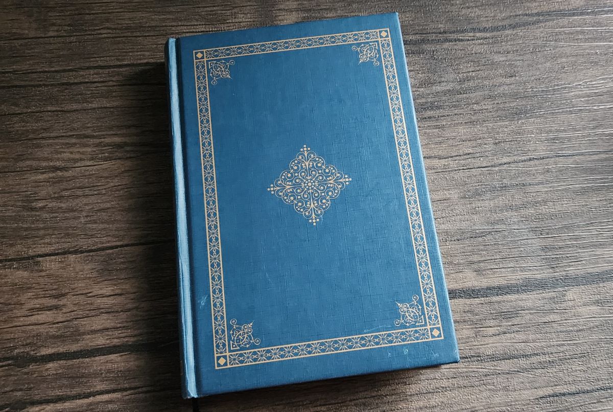 A hardcover book with a dark blue cover and intricate design of a frame with a diamond shape in the center in bronze gold foil on the cover.