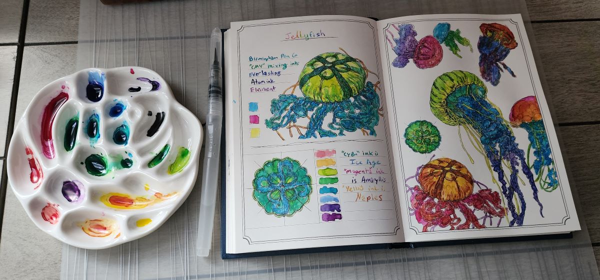 A ceramic paint palette with a rainbow of ink colors, water brush, and hardcover book open to a page of jellyfish painted in bright pink, orange, yellow, yellow green, green, blue green, blue, indigo, and purple that were all mixed from only three colors of ink: cyan, magenta, and yellow.