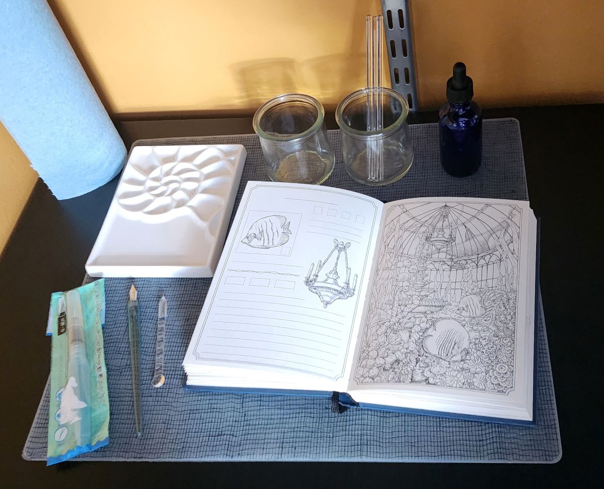 An example painting kit set up on a desk including a rectangle ceramic paint palette with a shell design, a water brush, a dip pen, a glass dip pen/muddler, the ink archiving book open to a unpainted page of a glass dome underwater, a small glass dropper bottle, and two small glass jars with a few glass stir sticks.