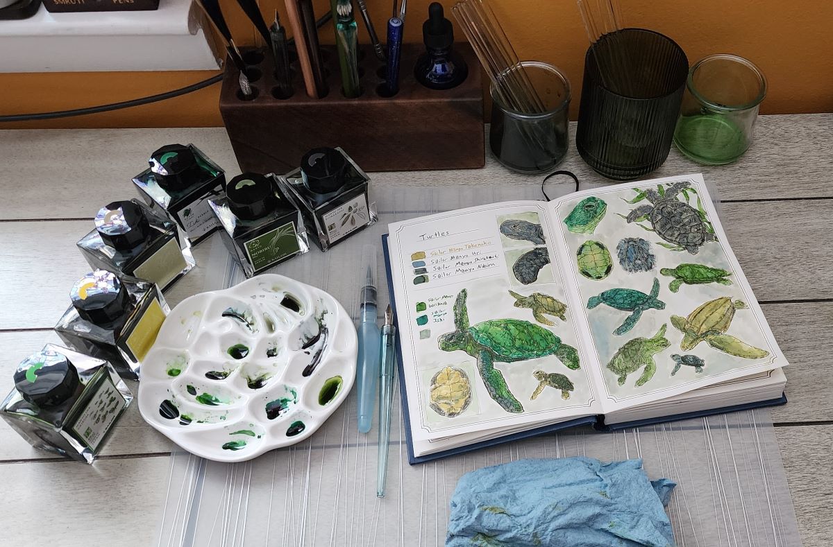 Fountain pen ink painting setup on desk with a silicone place mat, including several glass bottles of ink, a ceramic paint palette filled with drops of different shades of green ink, a water brush, a dip pen, a hardcover book opened to a page with drawings of turtles painted in shades of green, small glass jars with dirty ink water and glass stir sticks, a glass cup with clean glass stir sticks, a small dropper bottle filled with water, a blue shop towel.