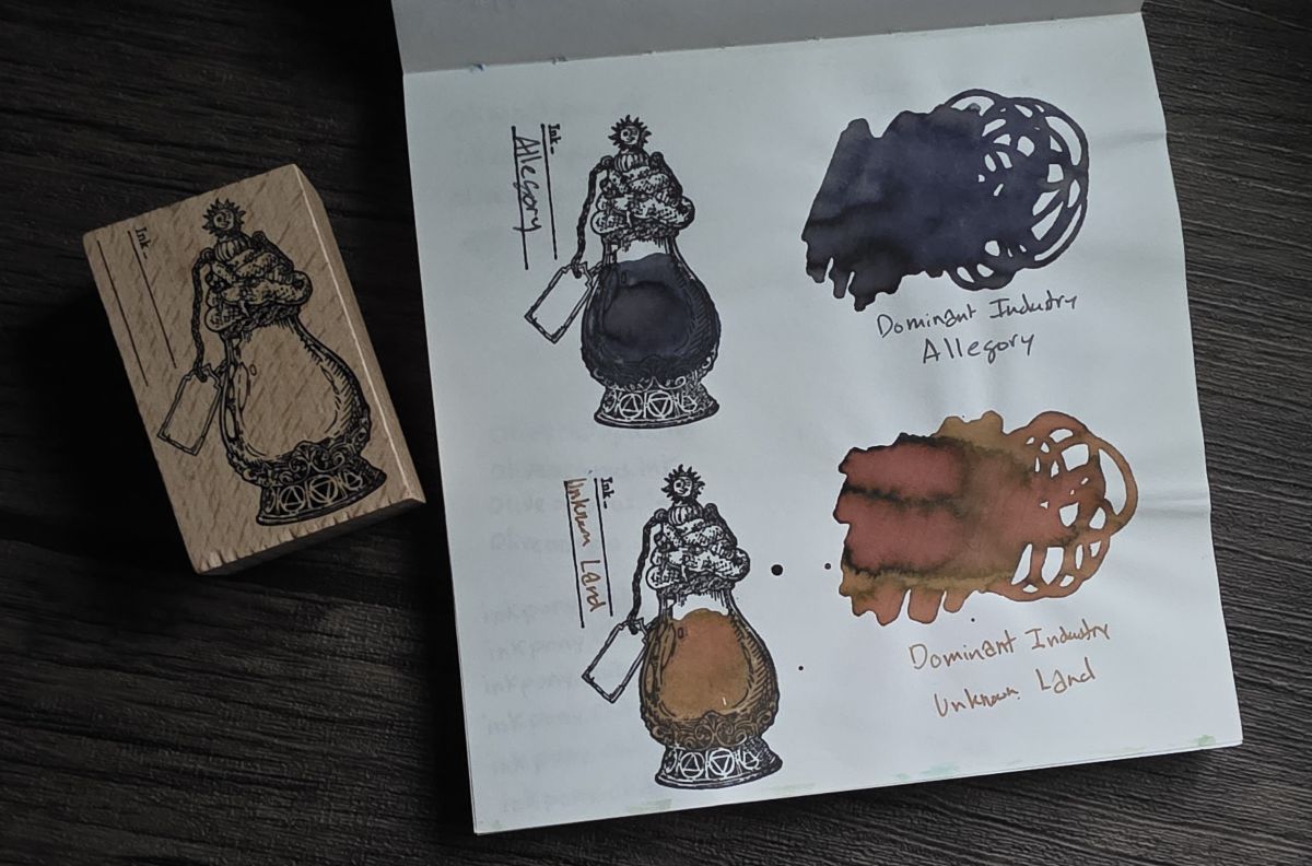 A square Midori Cotton notebook opened to a page with swatches of Dominant Industry Allegory, a dark blue and purple leaning gray, and Dominant Industry Unknown Land, a chromashading sepia with yellow tan and dusty pink shades. Next to the notebook is a wooden stamp featuring a shapely glass bottle with ornate base and cap, a small tag on a string around the neck, and two vertical lines with the word 'Ink' at the start of the first line.