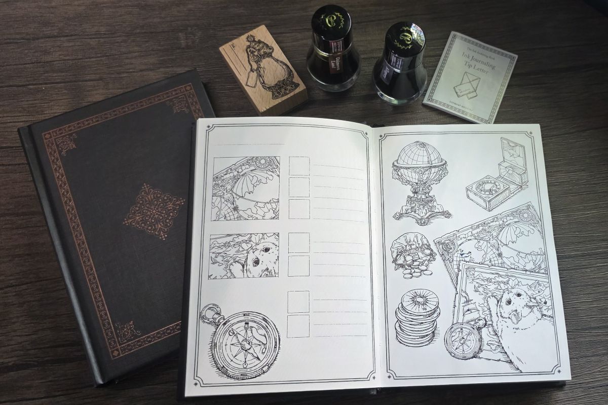 A hardcover B6 notebook with a black cover and bronze foil decoration, an open notebook with a black outline sketch of a globe, compass, coins, and a cat laying on a couple of maps. Next to the books are two glass bottles of fountain pen ink, a rubber stamp with a design of a fancy bottle, and a small paper pamphlet labeled Ink Journaling Tip Letter.