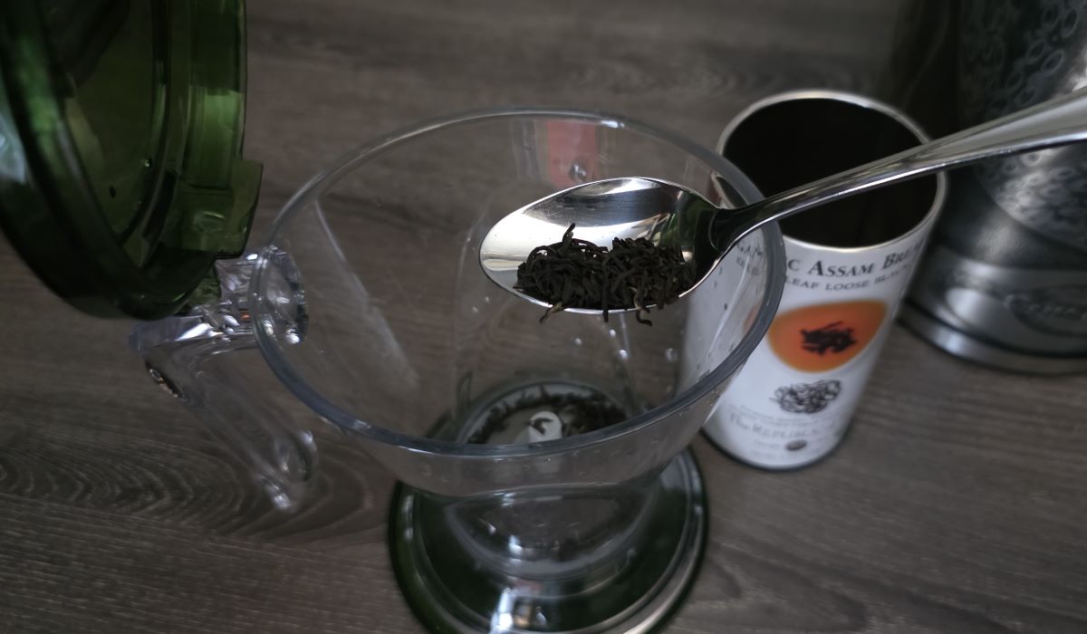 A teaspoon pouring loose leaf black tea into the Teaze tea infuser from a tall cylinder tin of tea. 