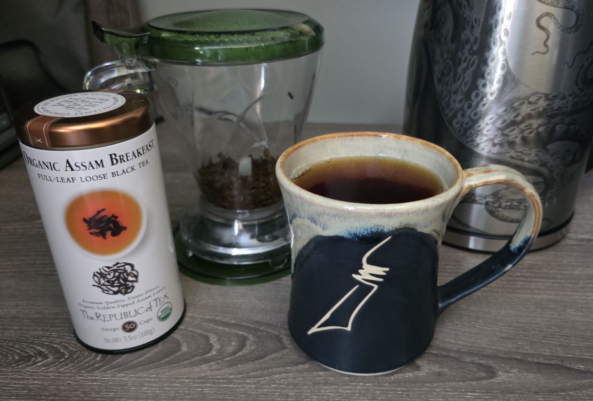 A ceramic handmade mug with nib design filled with steeped black tea, next to the Teaze tea infuser with the used loose-leaf tea, tin of tea, and electric kettle.