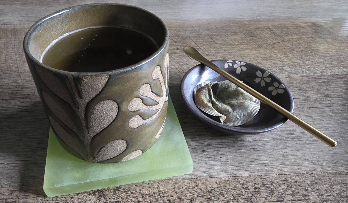 A handmade ceramic cup with olive green glaze and a textured gray leaf design filled with green tea, next to that is a small ceramic gray dish with white sakura flower design holding a used tea bag and small bronze color stirring spoon.