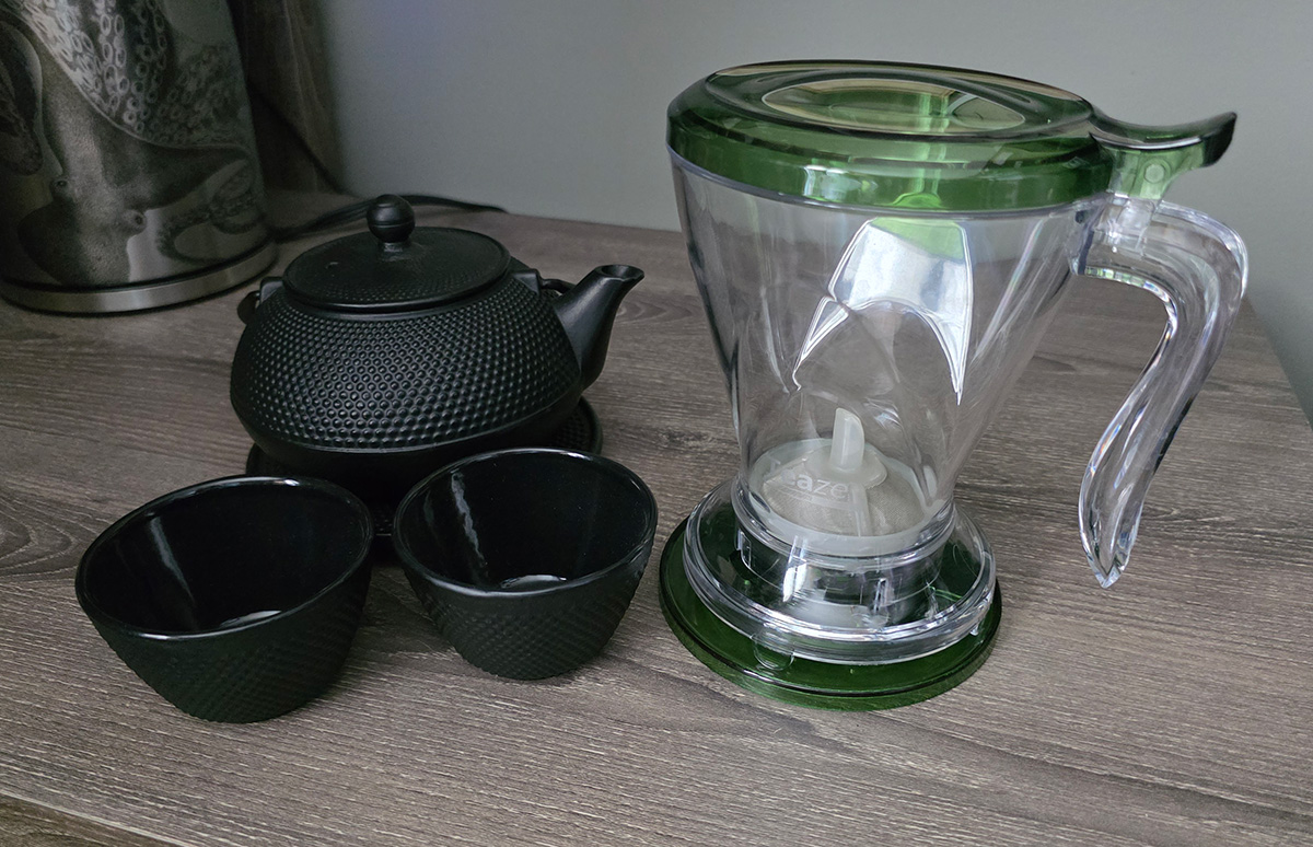 A small black cast iron teapot and two small cups, and a Teaze plastic tea infuser that holds almost two cups of tea and filters the steeped tea through the bottom into a mug or other container.