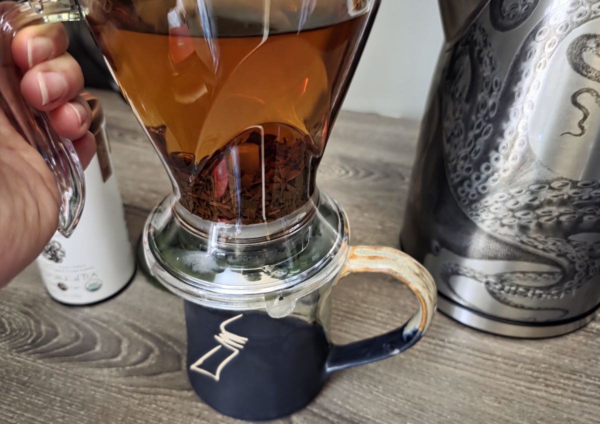 The Teaze tea infuser filled with steeped tea and loose tea leaves, sitting on top of a mug which has pressed up a plate on the bottom of the infuser to allow the tea to drain into the mug.