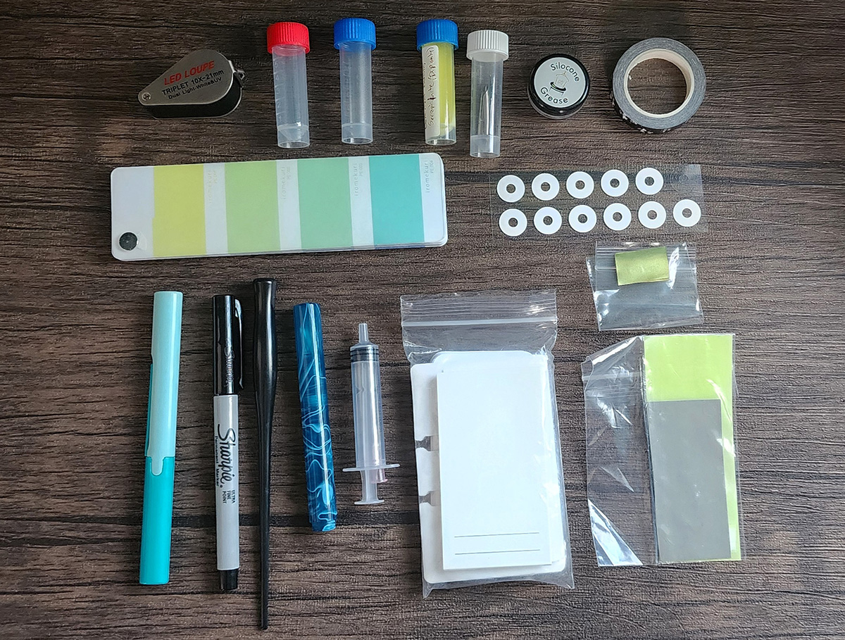 A 10x loupe, empty sample vials, sample vial with ink, sample vial with metal dip nibs, silicone grease, roll of narrow washi tape, stack of colorful, rectangle stickers, round ink vial labels, brass shims, pen-style scissors, black ultra fine point Sharpie, plastic nib holder, glass nib pen, blunt tip syringe, blank ink swatch cards, micro-mesh and mylar sheets.
