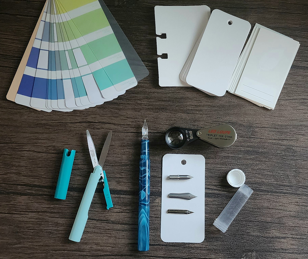 A colorful spread of green, blue, and purple rectangle stickers, a handful of ink swatch cards, compact pen-style scissors with a cap, a glass dip nib, a 10x loupe with light switch, three metal dip nibs.