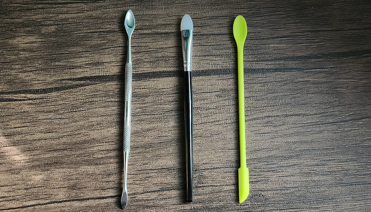 A metal tool with a small spoon shape at one end, a silicone makeup applicator with a plastic handle, and a green silicon tool with a thin spoon end and a mini spatula end.