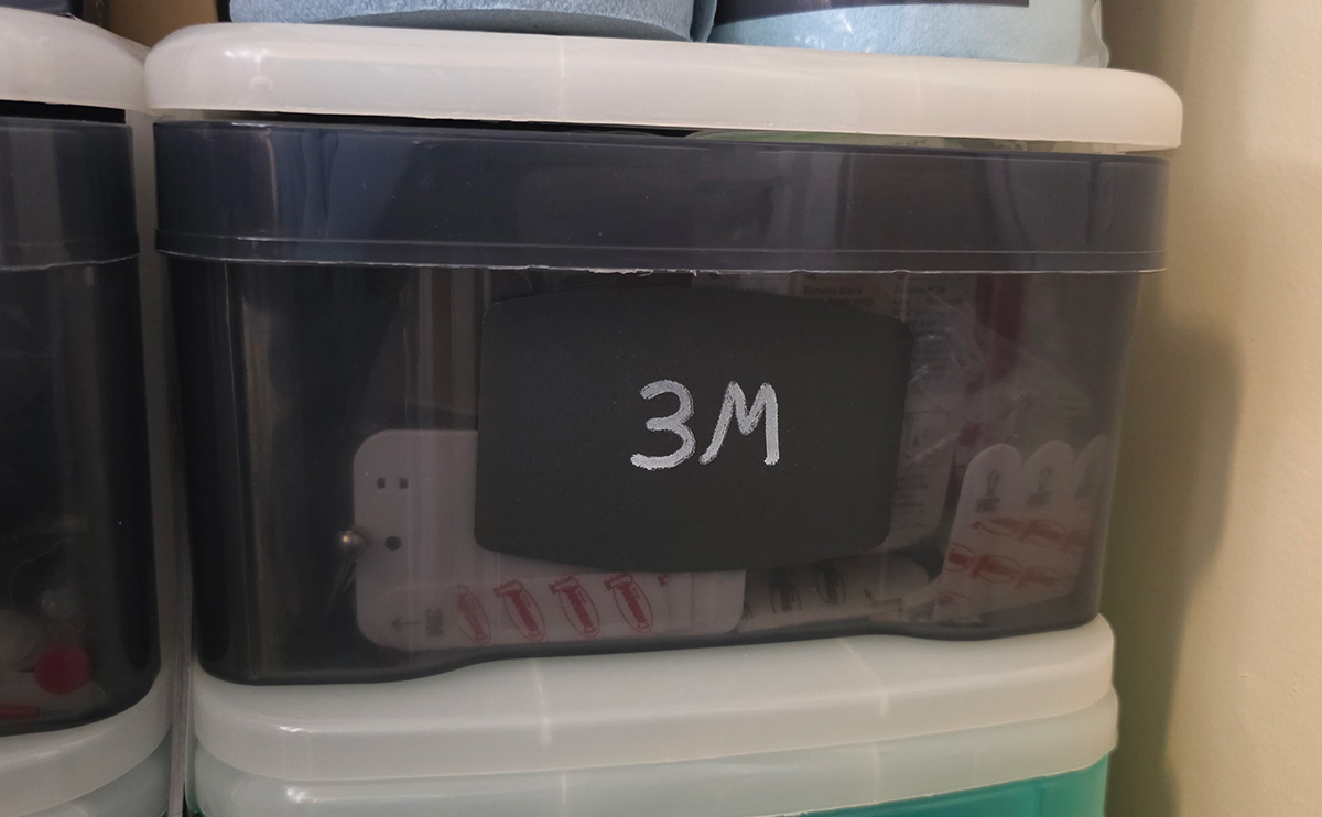 A gray plastic drawer on a shelf in a closet with a label on the front that says 3M