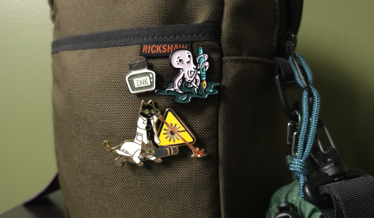 A dark green Rickshaw A5 field bag with 3 pins on the front pocket including a small green ink bottle, an octopus holding a fountain pen in a puddle of ink, and a pair of scientist cats using and playing with a laser.