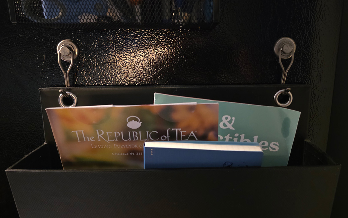 Two silver metal magnet hooks stuck to the side of a fridge holding a cardboard paper sorter with catalogs and a notebook inside.
