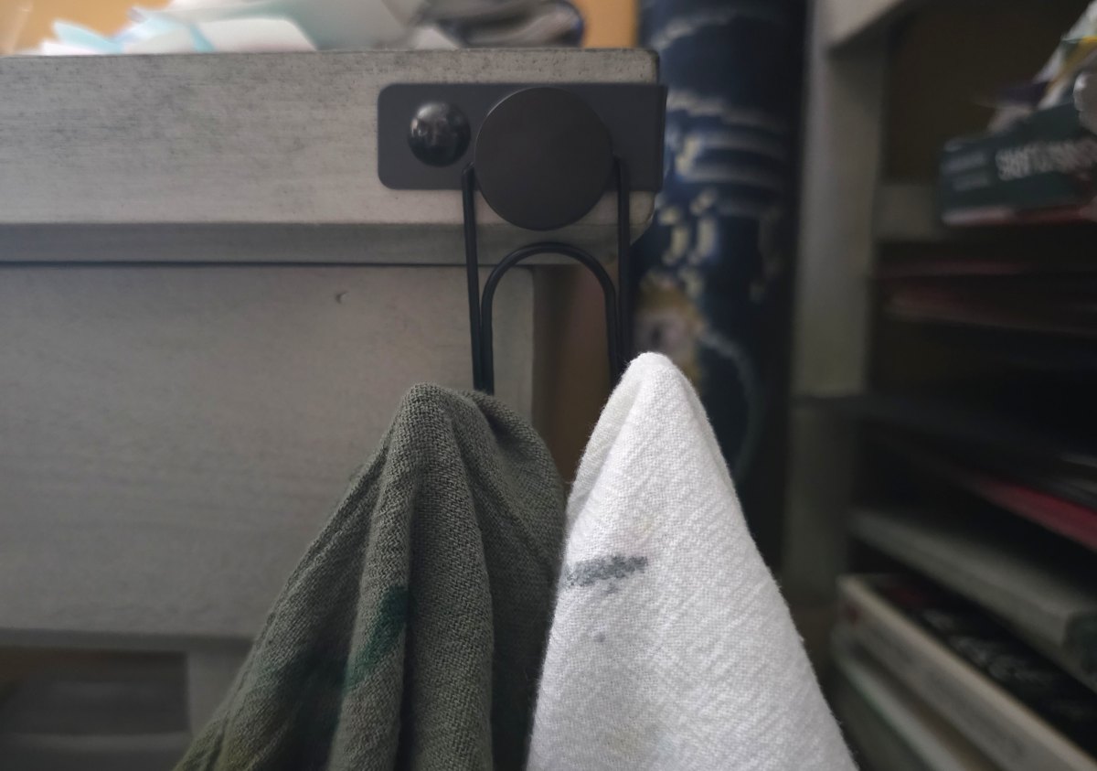 A black metal double hook attached to the corner of a desk with green and white ink-stained flour sack towels hanging on the hooks.
