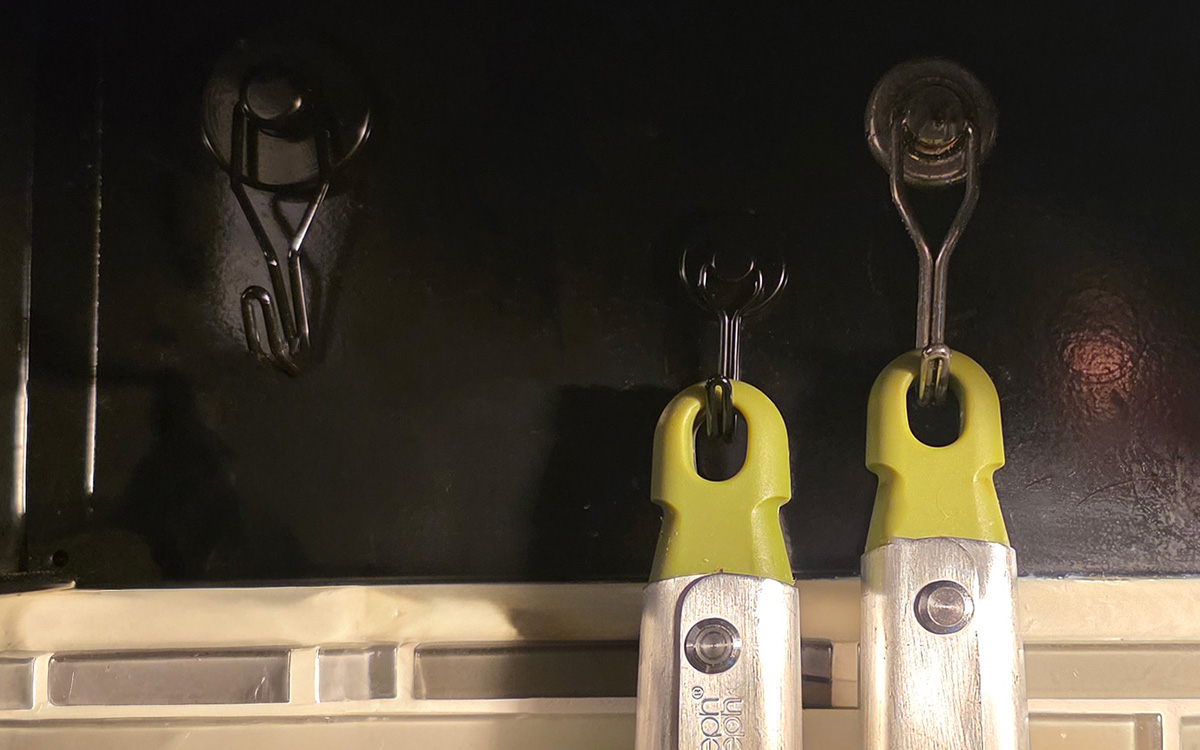 Three strong magnets with hooks attached by a hinge stuck to the back of a kitchen stove hood holding the handles of kitchen utensils.
