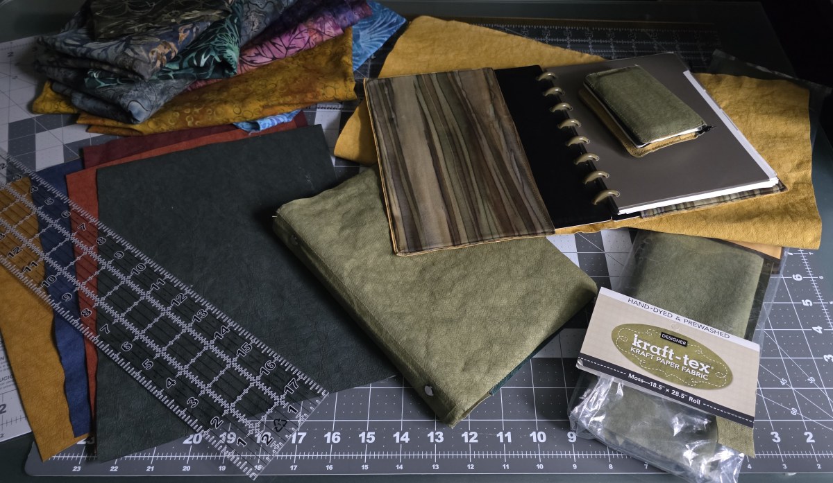 Kraft-Tex sewable paper material in several colors including pale yellow, dusty green, dark green, red, and blue, a stack of colorful batik fabrics in a variety of designs and colors, a prototype discbound cover with Kraft-Tex and fabric for the pockets, and prototypes of a small wallet and a 6 ring A5 binder made of Kraft-Tex.
