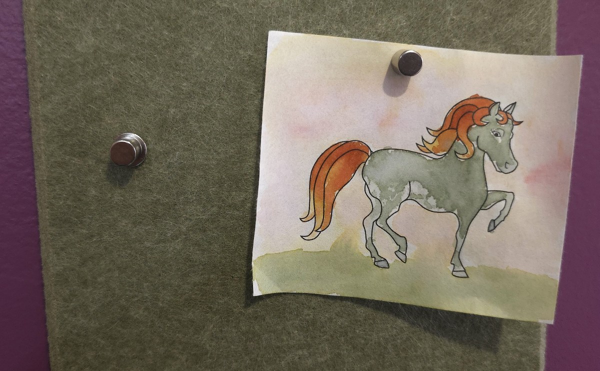 Two pins with a flat circular head and a magnet stuck on top in a green felt strip, one holding a small ink pony painting.
