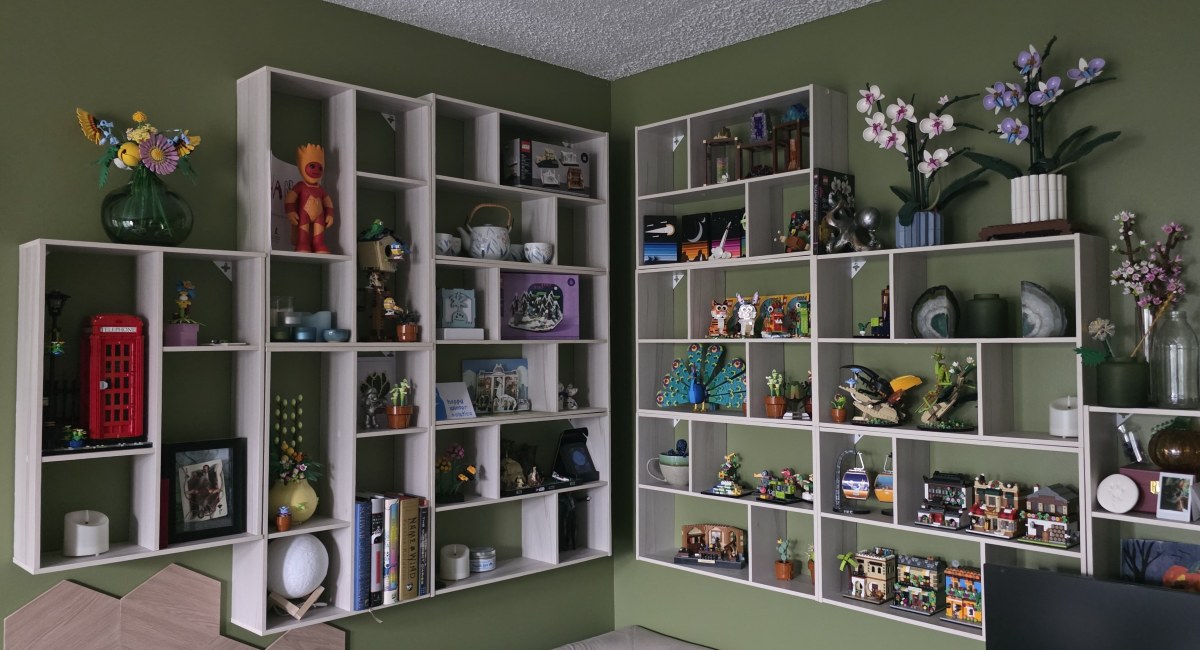 A dozen light gray rectangle wall shelves with square and rectangle cubbies arranged in the corner of the room and filled with Lego, books, lights, a tea set, and other knickknacks.
