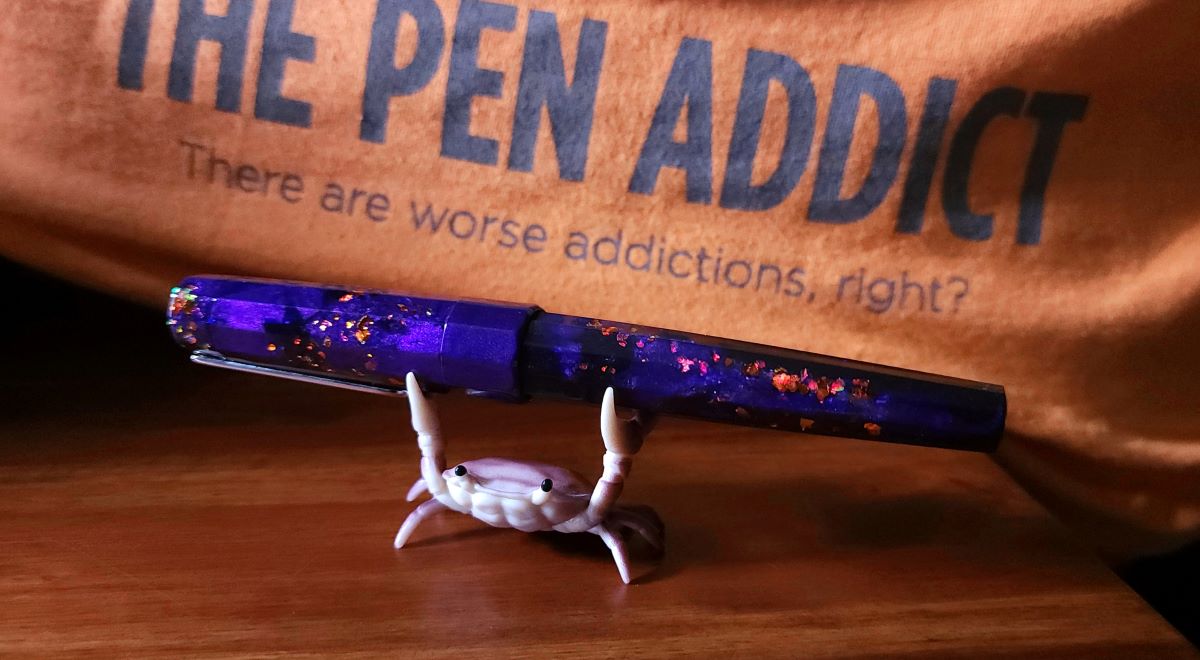 A purple resin pen with orange confetti and dark blue sparkles, held by a crab pen holder, in front of the Pen Addict logo and slogan.