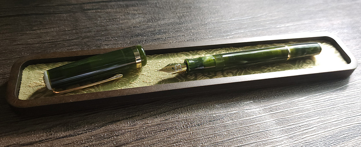 A slim, uncapped fountain pen with a chatoyant green material and gold-tone accents, clip, and nib.