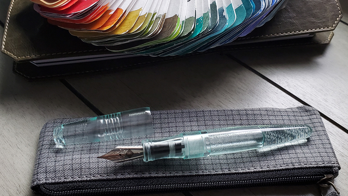 A tapered clear fountain pen that looks like slightly blue-green glass with a large reservoir for ink, resting on a pen pouch next to a spread of Col-o-ring ink swatches.