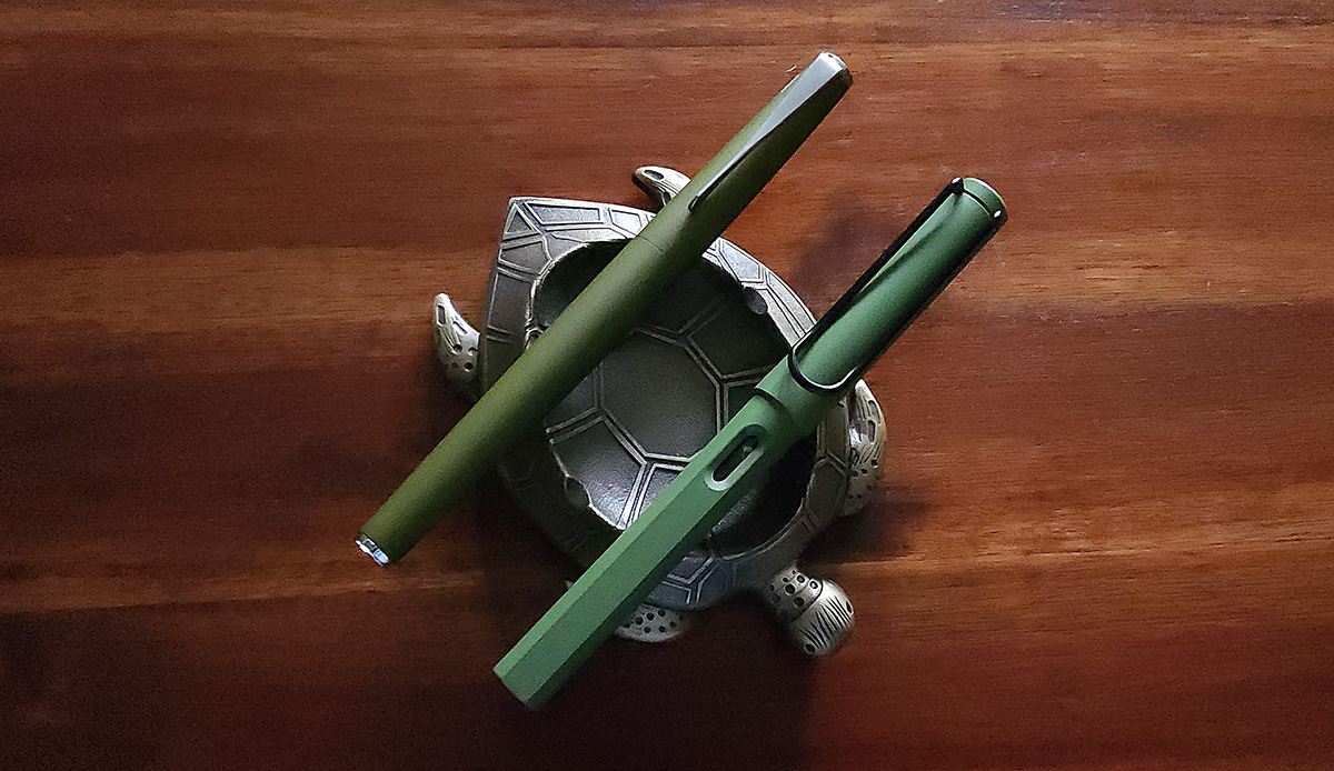 Two green fountain pens resting on a sea turtle pen holder, an olive green metal pen with a slim silver clip and a matte green plastic pen with a black wire clip and a small ink window.