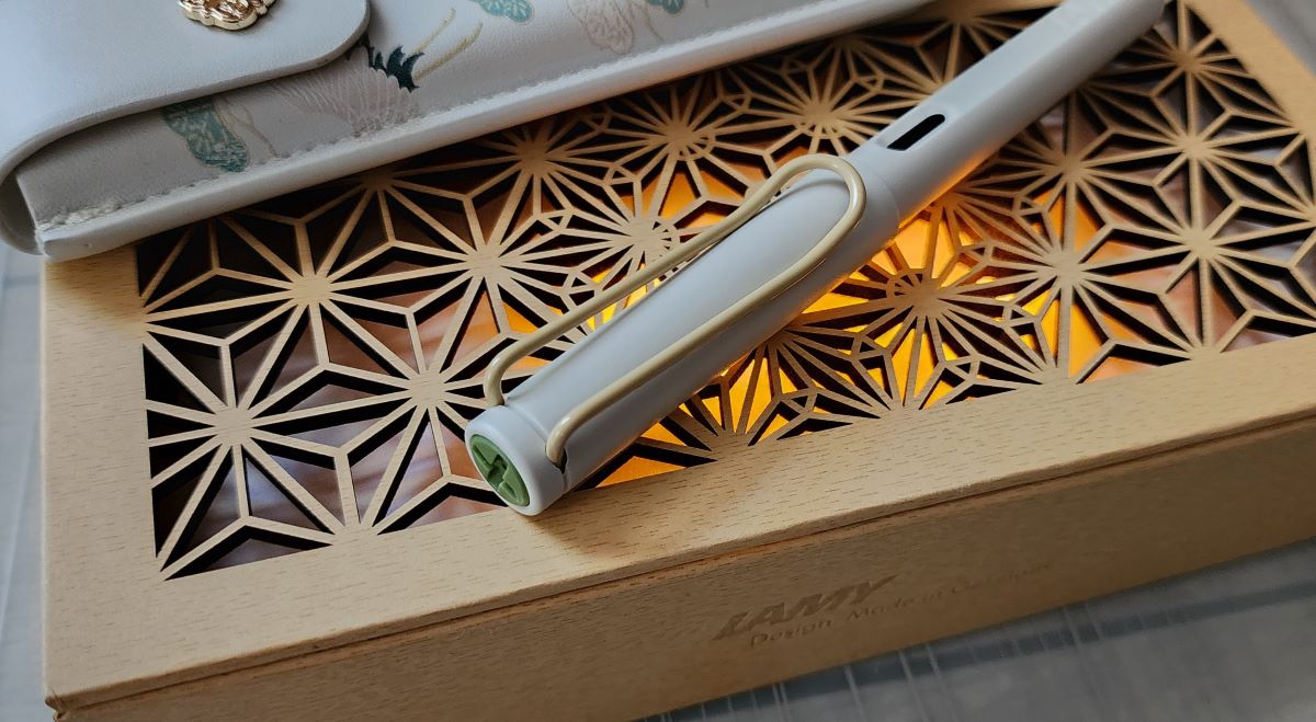 An off-white plastic fountain pen with a very light wood color wire clip and gray-green finial resting on a fancy carved box.