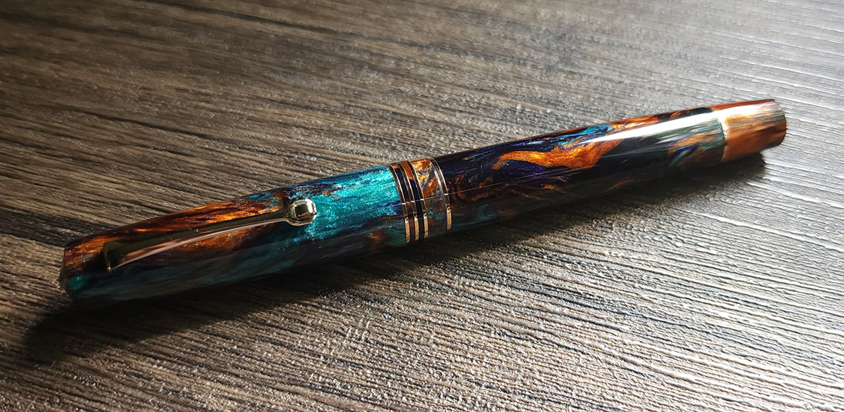 A fountain pen in a swirly orange, teal, and midnight purple pearlescent resin with thin rose gold accent bands and clip.