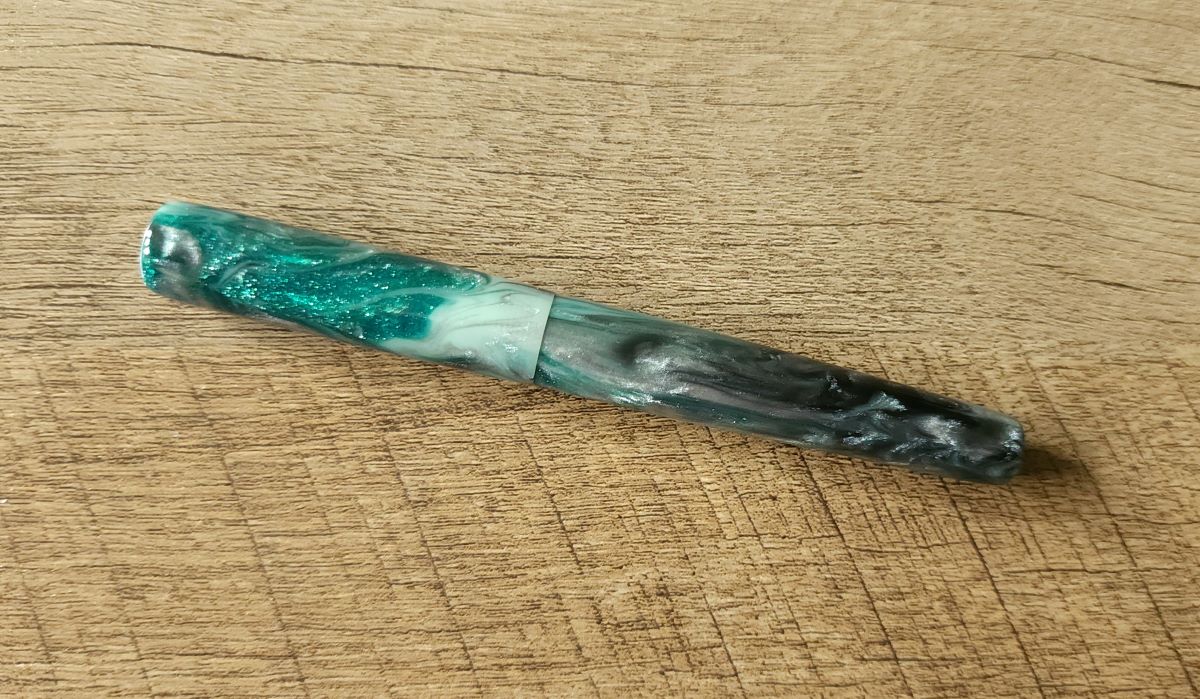 A hand turned fountain pen with a clean, slightly tapered shape in bright sparkly green, white, and dark silver swirled resin.