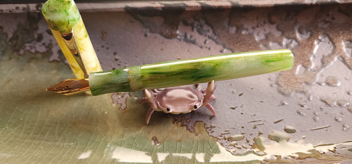 A turned resin fountain pen with gold tone nib and clip and translucent white with streaks of bright green and yellow green material, held by a small purple crab pen holder.