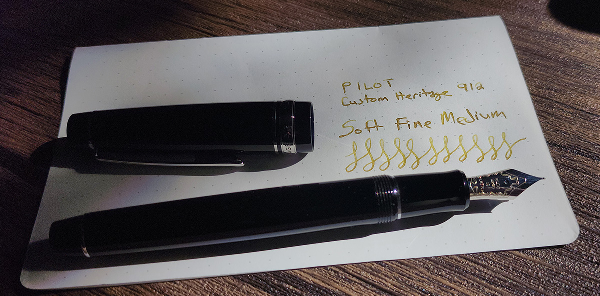 A black Pilot Custom Heritage 912 fountain pen with silver trim and flat ends with a writing sample of the Soft Fine Medium nib showing slight line variation.