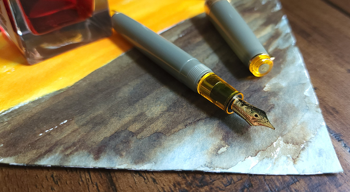 A Sailor Pro Gear Slim fountain pen with a gold nib, translucent yellow section, dusty gray green body and cap, and yellow finial with acorn design.